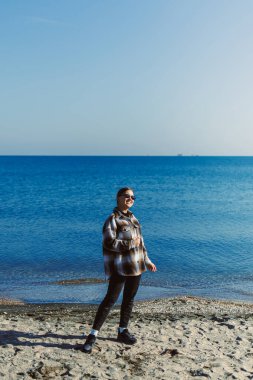Sahilde su kenarında güneş gözlüğü ve ekose gömlek takan bir adam var. Gökyüzü berrak ve deniz sakin dalgalarla.