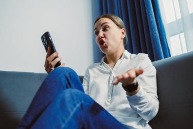 Uzun saçlı bir kadın oturma odasındaki bir kanepede oturur. O bir elinde bir telefon tutuyor ve konuşurken diğeriyle el kol hareketi yapıyor. Pencereden güneş ışığı akıyor..