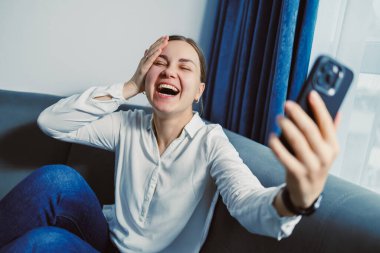 Bir kadın, yanındaki mavi perdeli bir kanepede oturuyor. Telefonunu tutuyor ve gülerken gülümsüyor. Güneş ışığı odayı dolduruyor, parlak bir atmosfer yaratıyor..