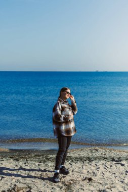 Bir kişi gün boyunca deniz kenarındaki kumlu plajda duruyor. Güneş gözlüğü ve kareli bir gömlek takıp denize bakıyorlar, manzaranın tadını çıkarıyorlar..