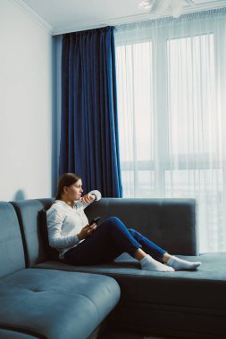 Geniş pencereli bir odada, koltukta oturan bir kadın. Elinde içki tutarken telefonuna bakıyor. Güneş ışığı perdelerin arasından gelir..