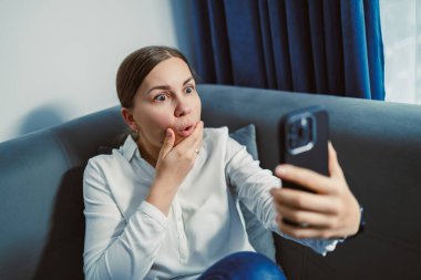 Bir kadın, elleri ağzında gri bir kanepede oturuyor. Oturma odasında içi doğal ışık dolu akıllı telefonuna bakarken şaşkın şaşkın bakıyor..