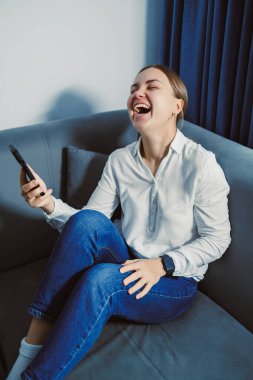 Bir kadın kanepede oturuyor, elinde akıllı bir telefonla gülüyor. Beyaz bir gömlek ve kot pantolon giyiyor. Akşam yaklaşırken pencereden gelen hafif ışıklandırma var..