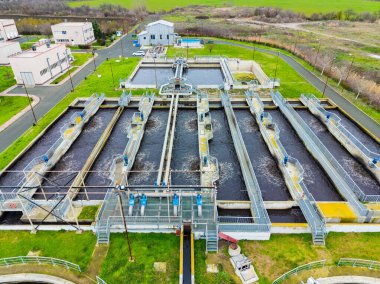 İşçiler suyu tedavi etmek için birden fazla tankın kullanıldığı bir atık su arıtma tesisi işletiyorlar. Tesis yeşil tarlaların ve binaların yakınında yer alıyor..