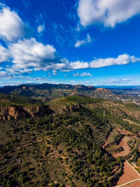 Geniş bir manzara, ağaç parçalarıyla dağları ve tarlaları yakalar. Gökyüzü bulutlarla aydınlık, ve manzara aşağıda tarım izleri gösteriyor, doğal bir manzara yaratıyor..