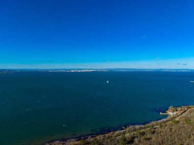 Sahne, denizde küçük bir tekneyle geniş bir okyanus manzarasını gösteriyor. Sahil şeridi uzaktaki binalarla görülebilir. Gökyüzü bulutsuz berrak.