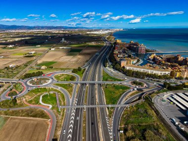 Manzara, sahile yakın tarlalar ve binalar boyunca uzanan bir otoyolu gösteriyor. Arka planda berrak gökyüzü ve mavi su görünür..
