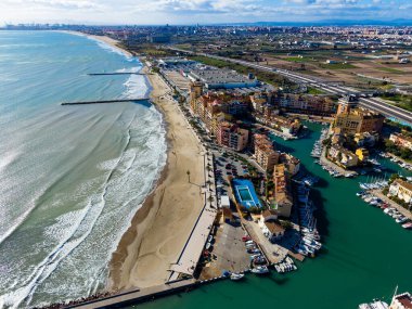 Sahne, sahili, binaları, marinası ve tekneleri olan bir sahil kasabasını gösteriyor. Arabalar arka planda yollarda giderken dalgalar kıyıya vuruyor..