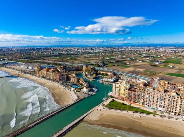 Sahne İspanya, Valencia 'daki deniz kıyısındaki kıyı binalarının görüntüsünü yakalıyor. Yeşil arazi kıyı boyunca görülebilir. Dalgalar kumsala çarpar açık mavi gökyüzünün altında.