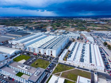 Birden fazla depo ve fabrikası olan büyük bir sanayi kompleksi. Çimenlik alanlar ve tarlalar binaları çevrelemektedir. Bulutlu bir gökyüzü manzarayı kaplıyor.