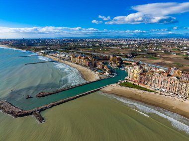 Kıyı şeridi boyunca uzanan binalar, kumlu sahilden geçen bir marinayla kesişiyor. Dalgalar kıyıya çarpar. Sahne, parlak mavi gökyüzünün altında bir su ve kara karışımını gösteriyor..