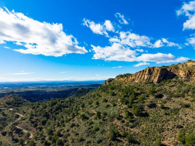 Parlak mavi gökyüzünün altında yeşil çalıları ve uzak dağları olan kayalık bir yamaç. Kabarık beyaz bulutlar yüzer manzaranın üzerinde.