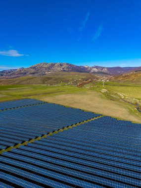 Güneş panelleri, dağların yakınındaki açık arazi boyunca uzanıyor. Gökyüzü açık ve mavi, birkaç bulutla birlikte. Tepeler ve ağaçlar arka planda görünür..