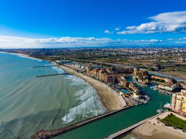 Uzaktan bakınca dalgalı bir sahil ve teknelerle dolu bir marina görünüyor. Binalar parlak mavi bir gökyüzünün altında bazı bulutlarla kıyı boyunca uzanır. Arazi mesafeye kadar uzanıyor..