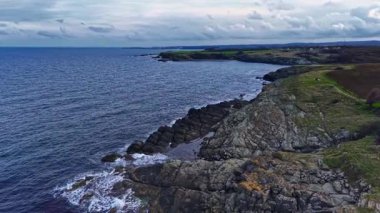 Şafakta, engebeli sahil kayalıkları yumuşak dalgaların üzerinde yükselir, karanlık kayalar suyun yüzeyini yararak bulutlu bir günde şaşırtıcı bir doğal manzara yaratır..