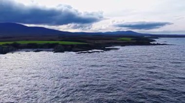 Kıyı boyunca sallanan otlar, kara bulutlar gökyüzünde belirdikçe, sakin sular ve alacakaranlıktaki uzak tepelerle çarpıcı bir kontrast oluşturur..