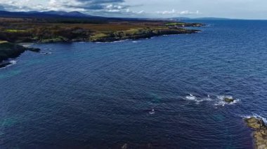 Dalgalar yavaşça kayalık kıyılara çarpar, suyun canlı mavilerini ve yeşilliklerini ortaya çıkarır. Geniş tepeler ve uzak dağlar bu huzurlu kıyı inzivasını oluşturur..