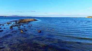 Sakin sular kayalara hafifçe çarpar, engin mavi gökyüzünü yansıtır. Uzakta, sakin bir kıyı şeridi görülebilir, alacakaranlıkta huzurlu bir tefekküre yol açar..