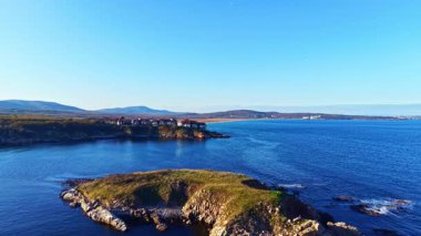 Sakin sular ufka doğru uzanırken kayalık kıyılar yumuşak dalgaları sallıyor. Yumuşak güneş ışığı manzarayı altın renklerde yıkar ve huzurlu bir atmosfer yaratır..