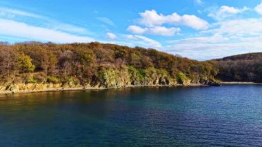 Canlı sonbahar yapraklarıyla süslenmiş sakin bir kıyı şeridi. Güneş tepemizde ışıl ışıl parlıyor. Kayalık kıyıya doğru süzülürken sakin suyu aydınlatıyor..