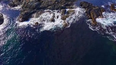 Titreşimli okyanus dalgaları sivri kayaların etrafında dans eder, derin mavi bir su zeminine köpüklü sıçramalar yaratır. Güneş ışığı yüzeyde parıldıyor, kıyı manzarasının dingin güzelliğini arttırıyor..