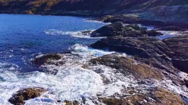 Parlak güneş ışığı, dalgaların engebeli kayalara çarpmasıyla sakin mavi sulardan yansıyarak doğal güzellik ve sükunetle dolu çarpıcı bir sahil sahnesi yaratır..