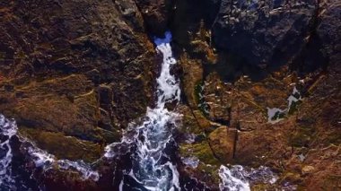 Dalgalar engebeli kıyı şeridine karşı enerjik bir şekilde dans eder, köpüklü beyaz armalar altın güneş ışığıyla vurgulanır. Kayalık arazi dokuları ve renkleri ortaya çıkararak büyüleyici bir kıyı manzarası yaratıyor..