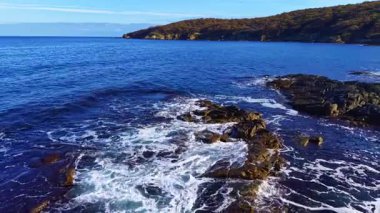 Güneş sakin bir sahil şeridinde yükseliyor, dalgaların ritmik bir şekilde çarpmasıyla karanlık kayaları aydınlatıyor, sakin bir denizin ve uzak tepelerin arasına beyaz köpük salıyor..