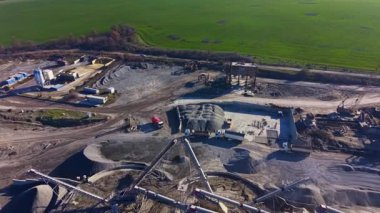 Ağır makineler, arka planda zengin yeşil tarlaları olan açık mavi bir gökyüzünün altında, kum ve çakıl yığınlarıyla çevrili inşaat sahasında özenle çalışıyor..