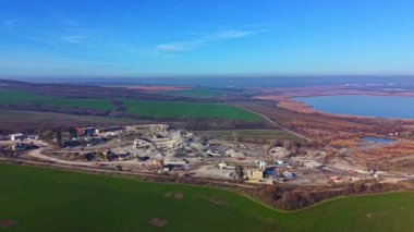 Hareketli yeşil tarlaların arasına yerleşmiş yoğun bir inşaat alanı, berrak gökyüzünü yansıtan huzurlu bir göl. İşçiler ve makineler çevreyi şekillendiriyor..