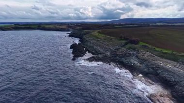Dalgalar yavaşça kayalık kıyı şeridine vururken bulutlar yukarı doğru sürükleniyor. Sahne, sakin bir akşamda, arka planda yuvarlanan tepelerle doğanın güzelliğini yakalar..