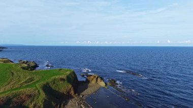 Dalgalar yavaşça kayalık sahil şeridine çarpar yemyeşil tepeler yakınlarda yükselerek doğanın gün ışığında parladığı huzurlu bir manzara yaratır..