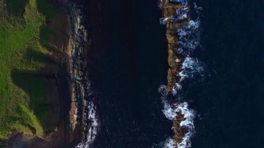 Sakin dalgalar engebeli kayalara çarparak derin mavi denizin üzerine güneş ışığını yansıtır. Kıyı şeridini çevreleyen yemyeşil çimenler gün ortasında sakin bir atmosfer yaratıyor..