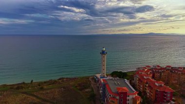 Güneş batarken, güzel bir sahil kasabası ılık renklerde güneşleniyor. Parıldayan denizin yanında duran büyüleyici bir deniz feneri ve kıyı şeridini benekleyen renkli binalar..