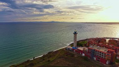 Deniz feneri, büyüleyici binalarla çevrili yumuşak dalgaları denetlerken, altın ışınlar kıyı şeridini aydınlatıyor. Manzara, dingin güzelliği ve ılık akşam parıltısıyla büyülüyor..