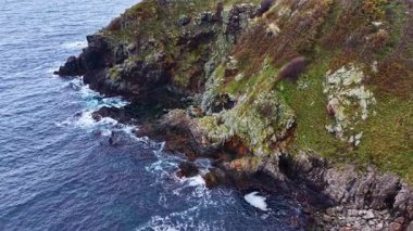Parlak mavi dalgalar verimli yeşilliklerle çevrili yosunlu kayalıklara nazikçe çarpar. Huzurlu sahne bir sükunet ve doğaya bağlılık hissi uyandırıyor..