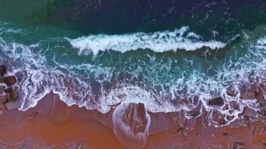 Dalgalar kıyıya doğru hareket ederek, kuma kuvvetle vuruyor. Su kayaların üzerinde köpük oluşturur. Sahne, parlak güneş ışığı altında açık bir günde sahili gösteriyor..