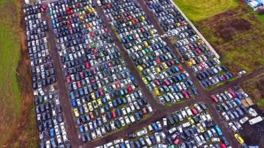 Hava görüntüsü, park halindeki birçok farklı renk ve boyutta arabalarla dolu büyük bir otoparkı gösteriyor. Yakınlarda, bazı kirli yolların görüldüğü çimenlik bir alan var..