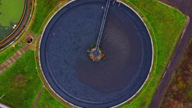 Yukarıdan bakıldığında geniş dairesel tankları olan bir su arıtma tesisi görülüyor. Tanklar karanlık suyla dolu ve bir tank boyunca uzanan bir platform var. Tesisi çevreleyen yeşil alanlar.