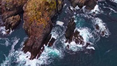 Dalgalar gün batımında sert kayalıklara vururken okyanus yumuşak mavi ve beyaz renkleri yansıtıyor. Bu sahne, karayla su arasındaki uyumu, huzurlu bir ortamda vurgular..