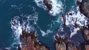 Turkuaz sular kayalık kıyılarla kaynaşır ve dalgaların kayalıklara çarpmasıyla çarpıcı bir kontrastlık yaratır. Deniz yukarıdaki canlı gökyüzünü yansıtır..