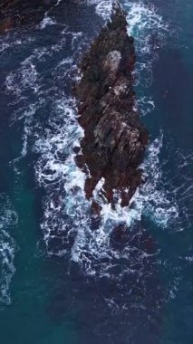 Dalgalar parlak mavi okyanusta beyaz köpük oluşturan pürüzlü bir kaya oluşumuna çarpar. Güneş suyun üzerinde parıldıyor, nefes kesici bir kıyı manzarası sergiliyor..