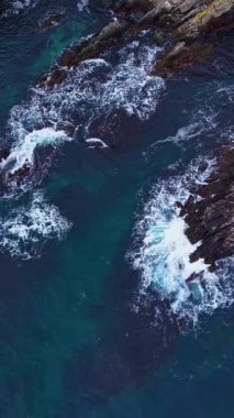 Parlak dalgalar engebeli kayalara ritmik olarak çarpar, suyu mavi ve yeşil tonlarıyla boyarken, güneş huzurlu bir sahil şeridinde batmaya başlar..