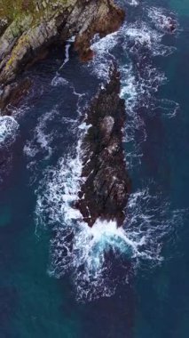 Dalgalar, güzel bir sahil şeridi boyunca uzanan pürüzlü kayalara güçlü bir şekilde çarpar. Canlı mavi su, günbatımı renklerinin ipuçlarıyla çelişir. Nefes kesici ve sakin bir atmosfer yaratır..