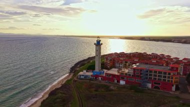 Batan güneş, su kenarındaki canlı binalarla çevrili bir sahil fenerinin üzerine sıcak bir ışık saçıyor. Huzurlu ve resimli bir manzara yaratıyor..