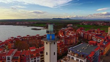 Uzun bir deniz feneri, parlak suların ve temiz bir gökyüzünün altındaki yeşil tarlaların üzerine kurulmuş kırmızı çatılı binaları olan canlı bir kasabaya bakıyor. Sahne sıcaklık ve cazibe saçıyor..