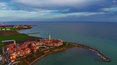 Parlak gün batımı, sakin bir sahil kasabasının üzerine sıcak renkler salıyor. Sahil şeridinde uzun bir deniz feneri duruyor. Alacakaranlıkta huzurlu bir atmosfer yaratıyor..