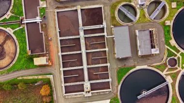 Kentsel su arıtma merkezinin üstünde dairesel tanklar ve uzun dikdörtgen kanallar atık suyu temizlemek ve geri dönüştürmek için karmaşık sistemi gösteriyor. Yeşil peyzaj sanayi sahnesine zıt düşüyor.