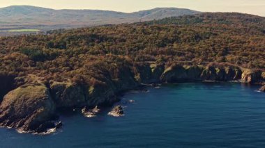 Yumuşak dalgalar kayalık kıyılara vururken, bu sakin sahil şeridinin altın saatindeki güzelliğini vurgulayan bereketli yeşil kayalıklar dramatik kayalıkları sarar..