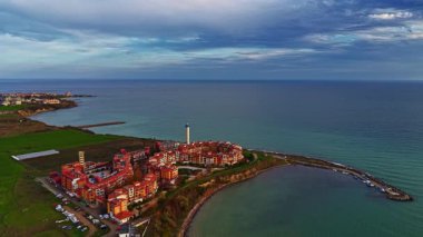 Çarpıcı bir sahil manzarası, yemyeşil sularla çevrili büyüleyici bir sahil köyünü yakalıyor. Gökyüzü mavi ve turuncu tonlarda boyanır alacakaranlık çökerken.